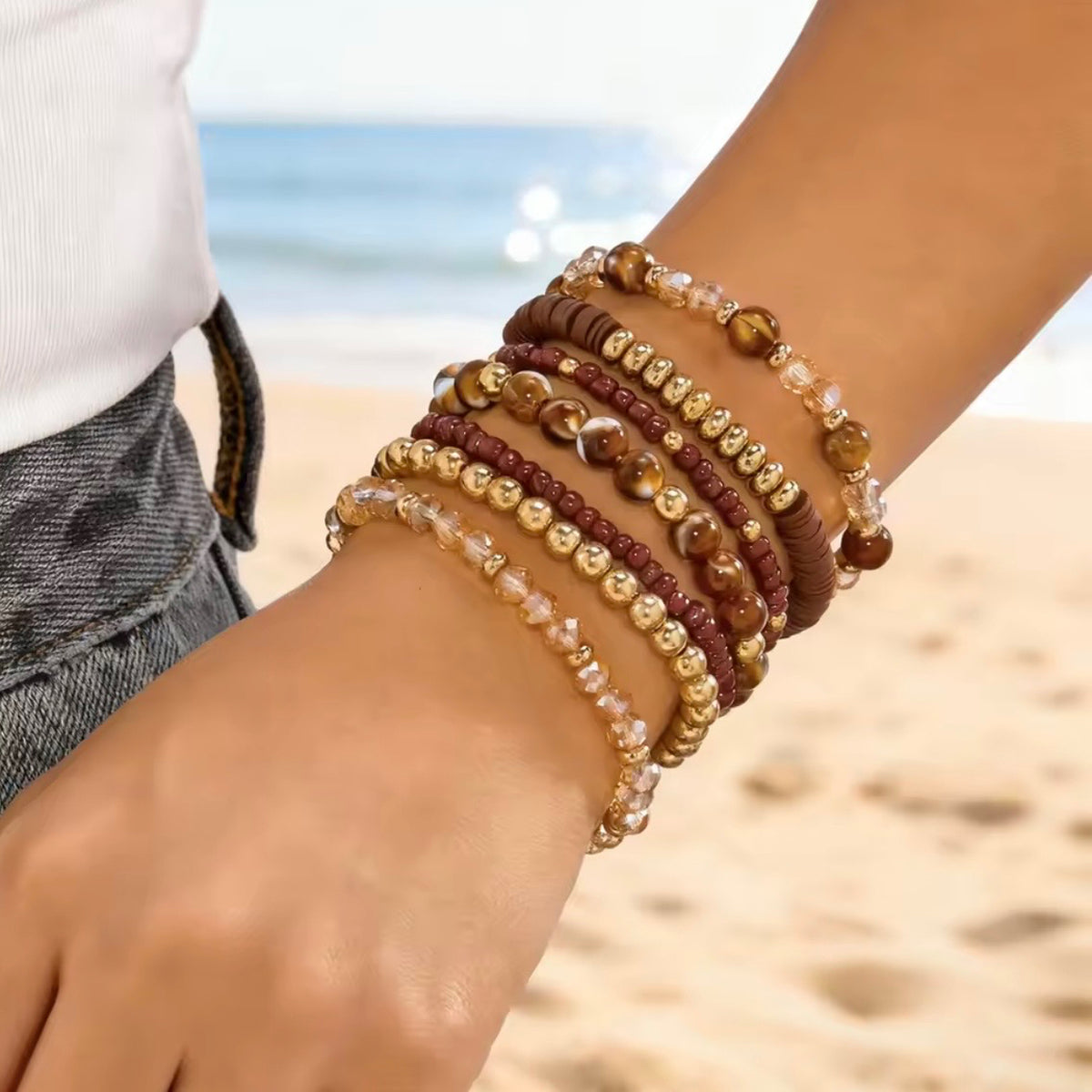 Brown and gold boho beaded bracelets worn as a layered stack on a wrist against a softly blurred beach background.