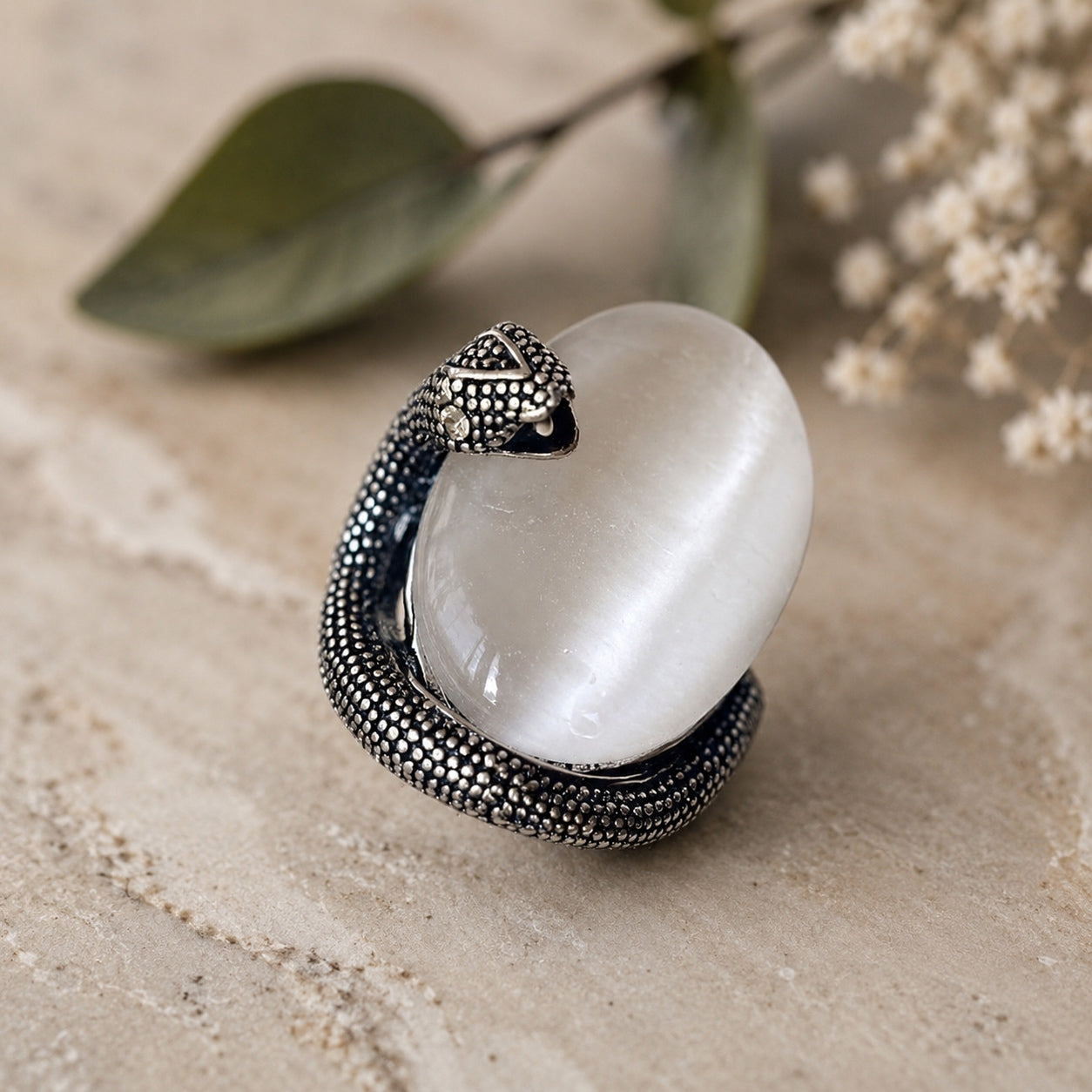 Cat’s Eye Stone Snake Ring with an oval white cat’s eye gemstone displayed on a natural stone surface with botanical accents.