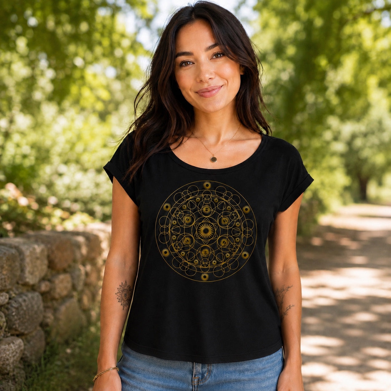 Woman wearing the Geometric Mandala Roll Cuff Tee in black featuring an intricate gold geometric mandala design, styled outdoors on a sunny nature path.
