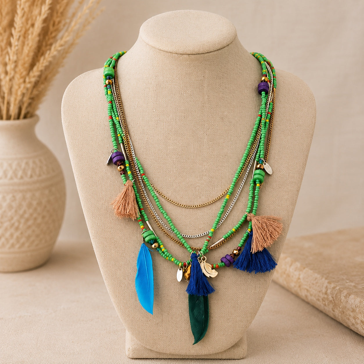 Green layered boho beaded necklace with blue and green feather pendants, tassels and mixed metal chains displayed on a linen jewelry bust.