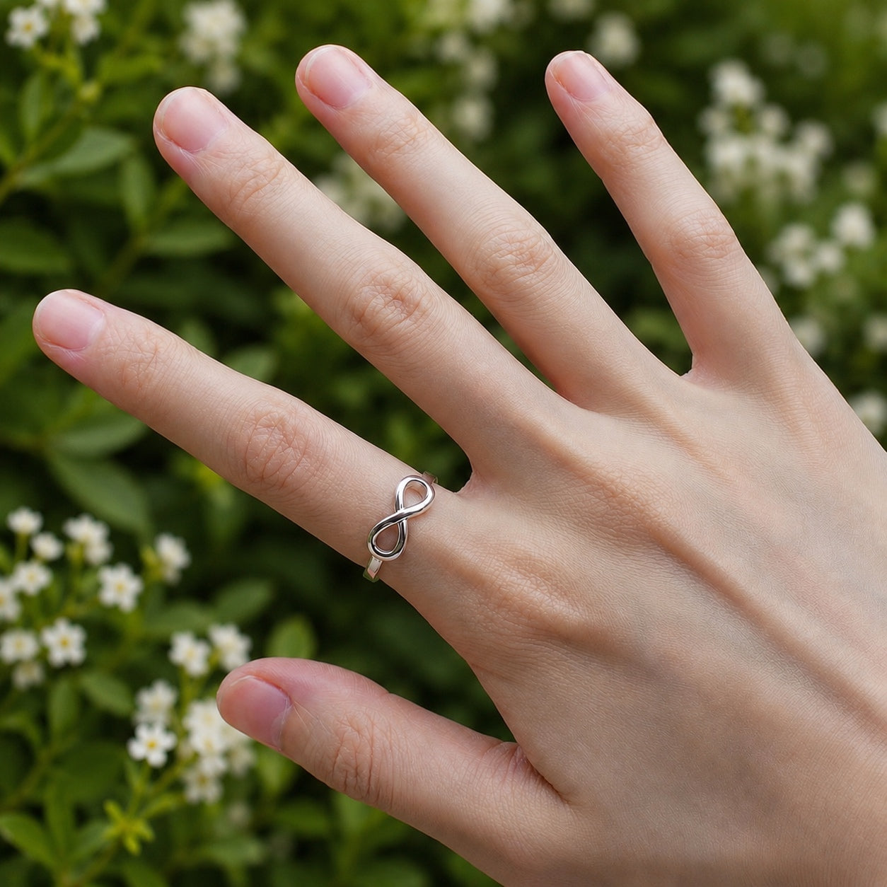 Infinity Ring in polished sterling silver worn on a woman’s hand with a softly blurred floral garden background.
