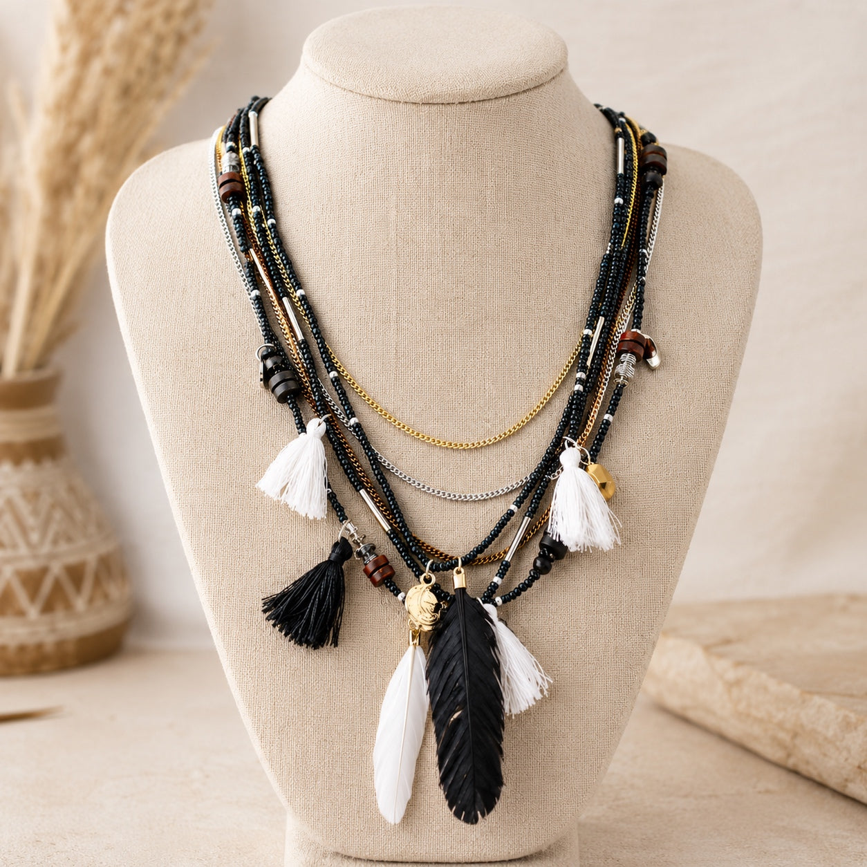 Black and white layered boho necklace with feather pendants, tassels and mixed metal chains displayed on a linen jewelry bust.