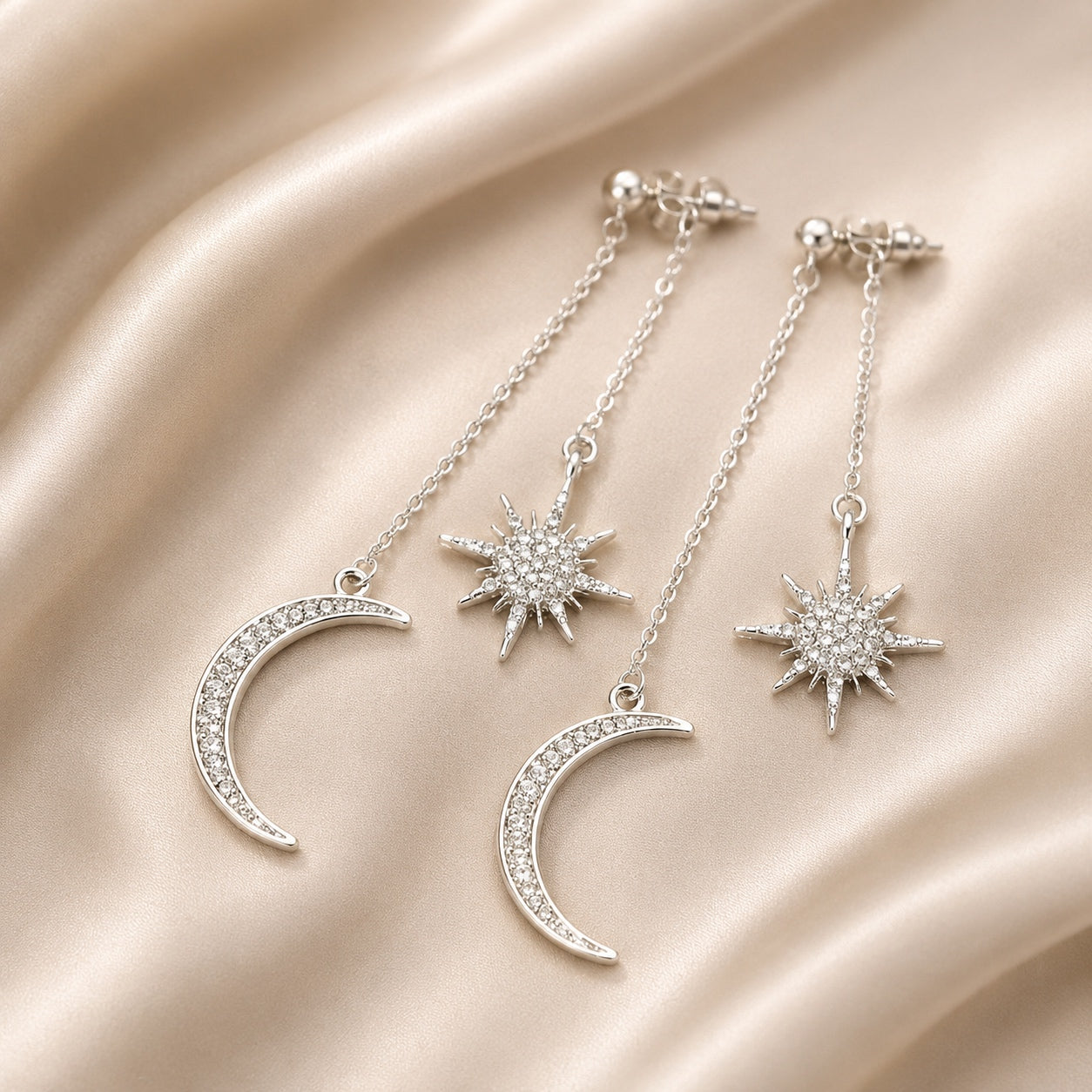 Silver Celestial Dangle Earrings with crystal moon and star charms displayed on luxurious champagne satin fabric.
