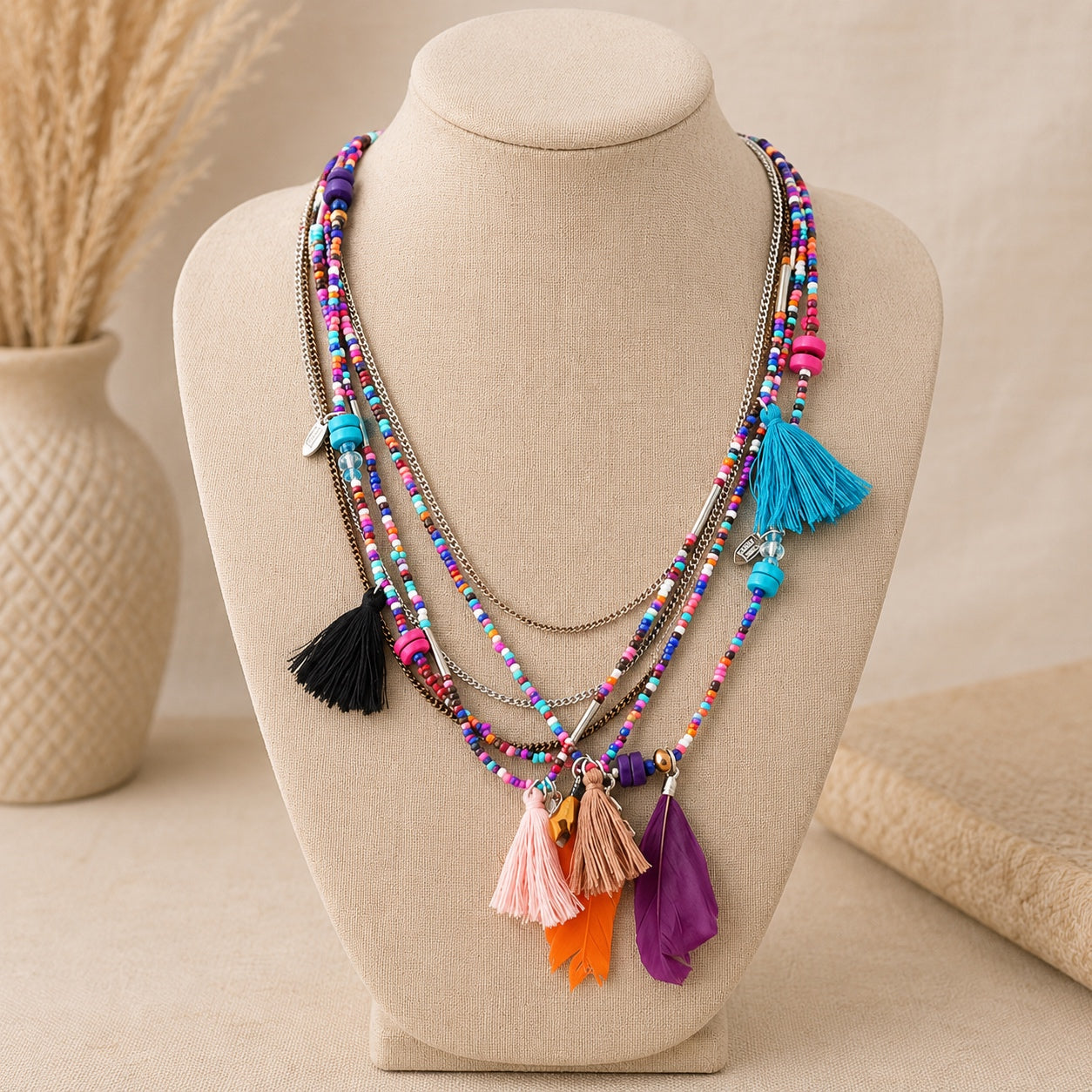 Colorful layered boho beaded necklace with tassels, feather pendants and mixed metal chains displayed on a linen jewelry bust.
