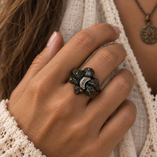 A close-up of a hand wearing a textured, organic-shaped ring with a dark green patina and a silver crescent moon in the center, styled with a cream knit sweater.