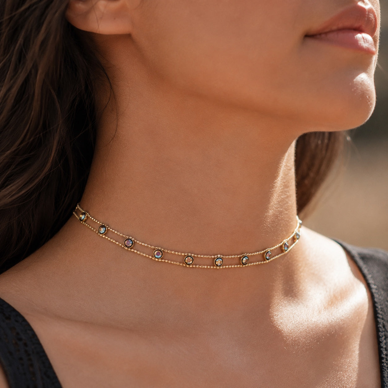 Woman wearing the Dainty Gems Choker Necklace featuring delicate double gold chains with shimmering iridescent crystal accents in a close-up lifestyle portrait.