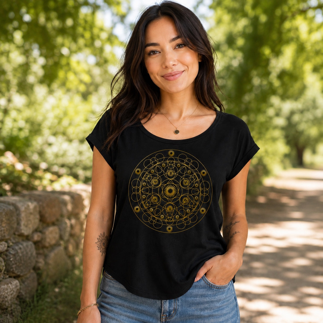 Woman wearing the Geometric Mandala Roll Cuff Tee in black featuring an intricate gold geometric mandala design, styled outdoors on a sunny nature path.