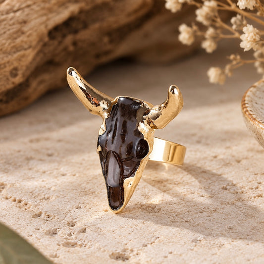 Gold-tone bull skull statement ring with dark marbled detailing on a beige stone surface.