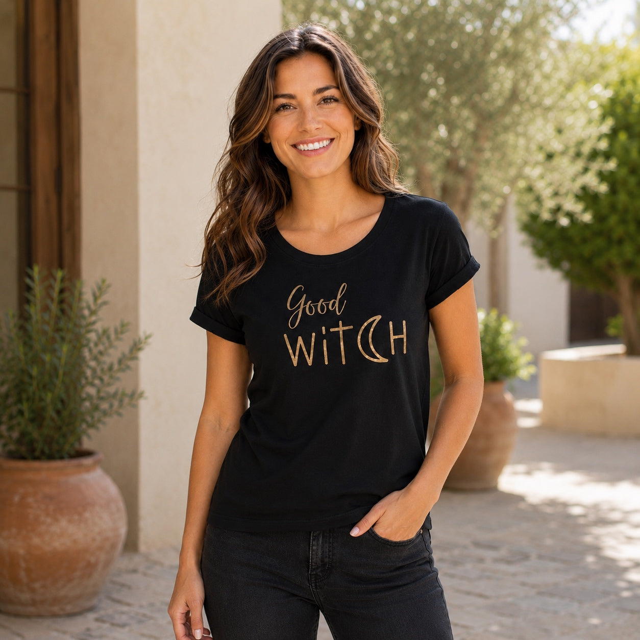Woman wearing the Good Witch T-Shirt in black with metallic gold lettering, smiling outdoors in a sunny courtyard.