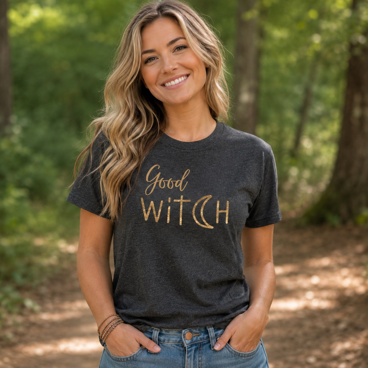 Woman wearing the Good Witch T-Shirt in dark heather gray with metallic gold lettering while standing on a wooded trail.