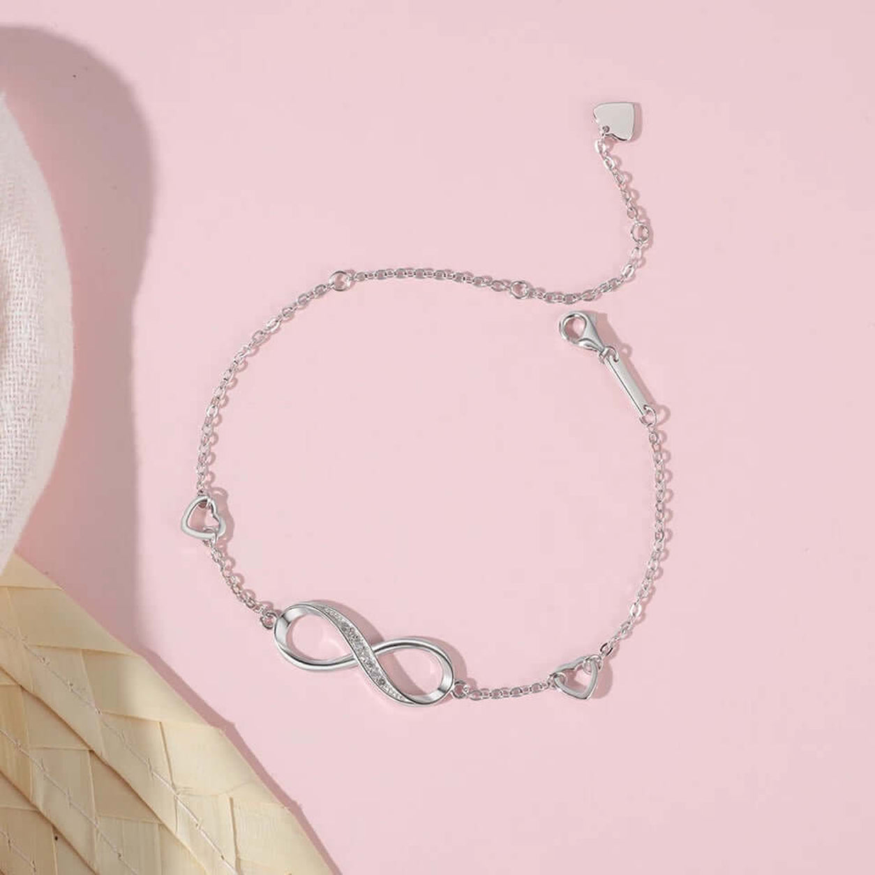 Flat lay of the Infinity Bracelet on a soft pink background showcasing the infinity charm, crystal detail, and adjustable heart chain.