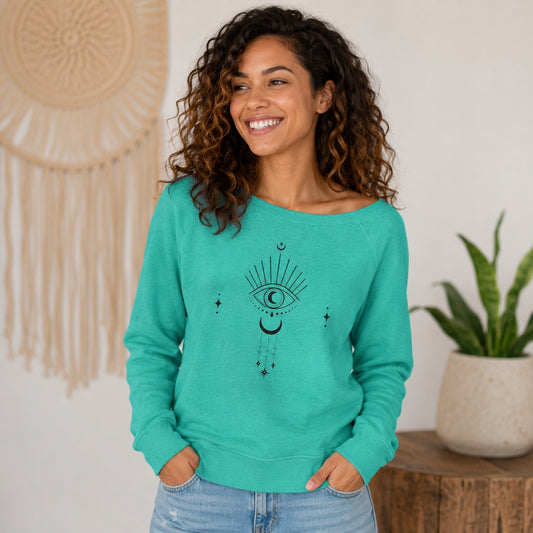 Woman wearing the teal Moongazer Wide Neck Sweatshirt with celestial eye and moon graphic in a cozy boho-inspired home.