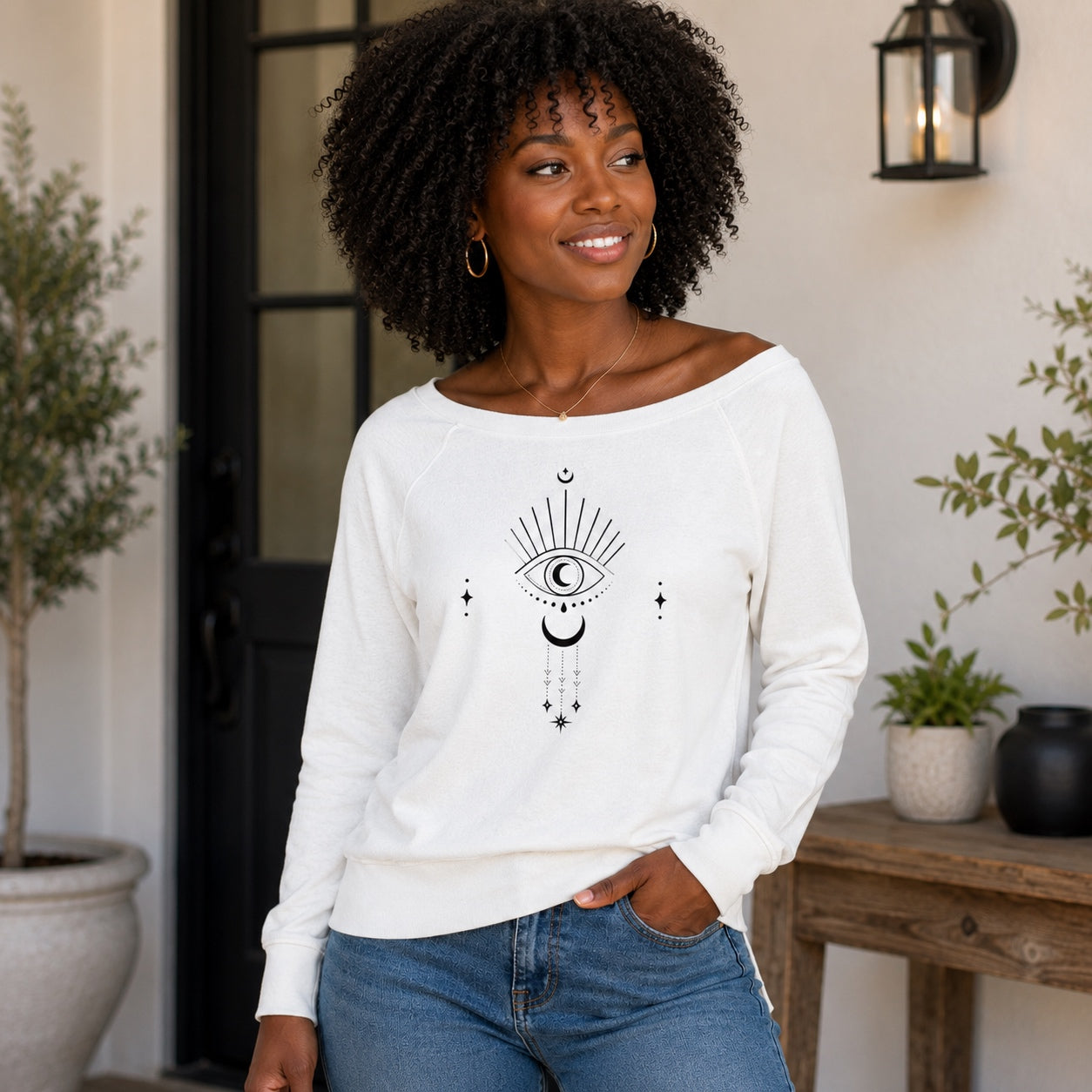 Woman wearing the white Moongazer Wide Neck Sweatshirt with celestial moon and eye graphic in a modern outdoor setting.
