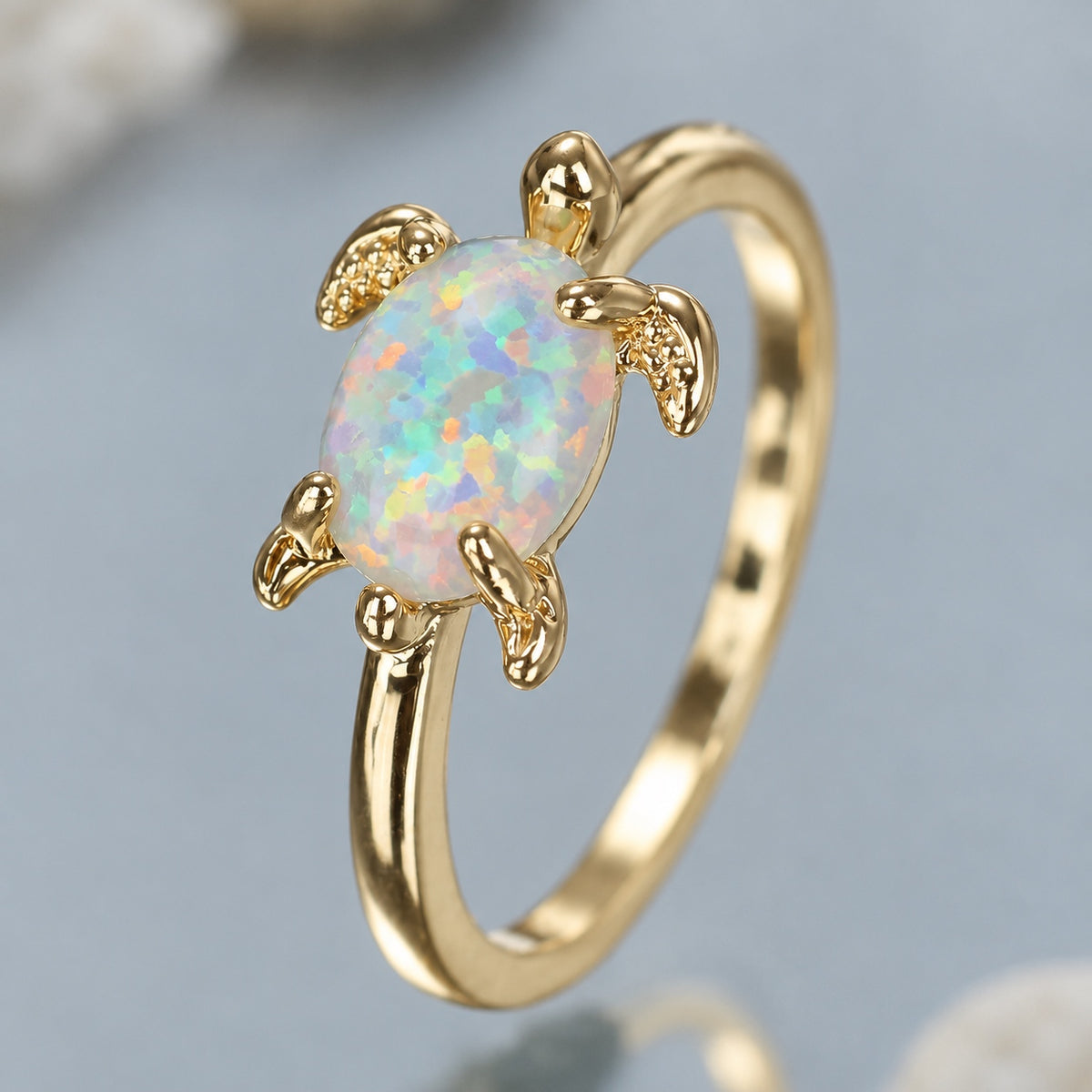A gold ring shaped like a sea turtle, featuring an oval opal gemstone with a multicolored iridescent play-of-color effect, set against a soft light blue-gray background.