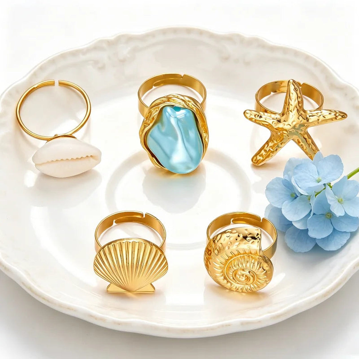 Seaside Treasures Ring Set displayed on a decorative plate featuring gold-tone starfish, seashell, cowrie shell, spiral shell, and aqua statement rings.