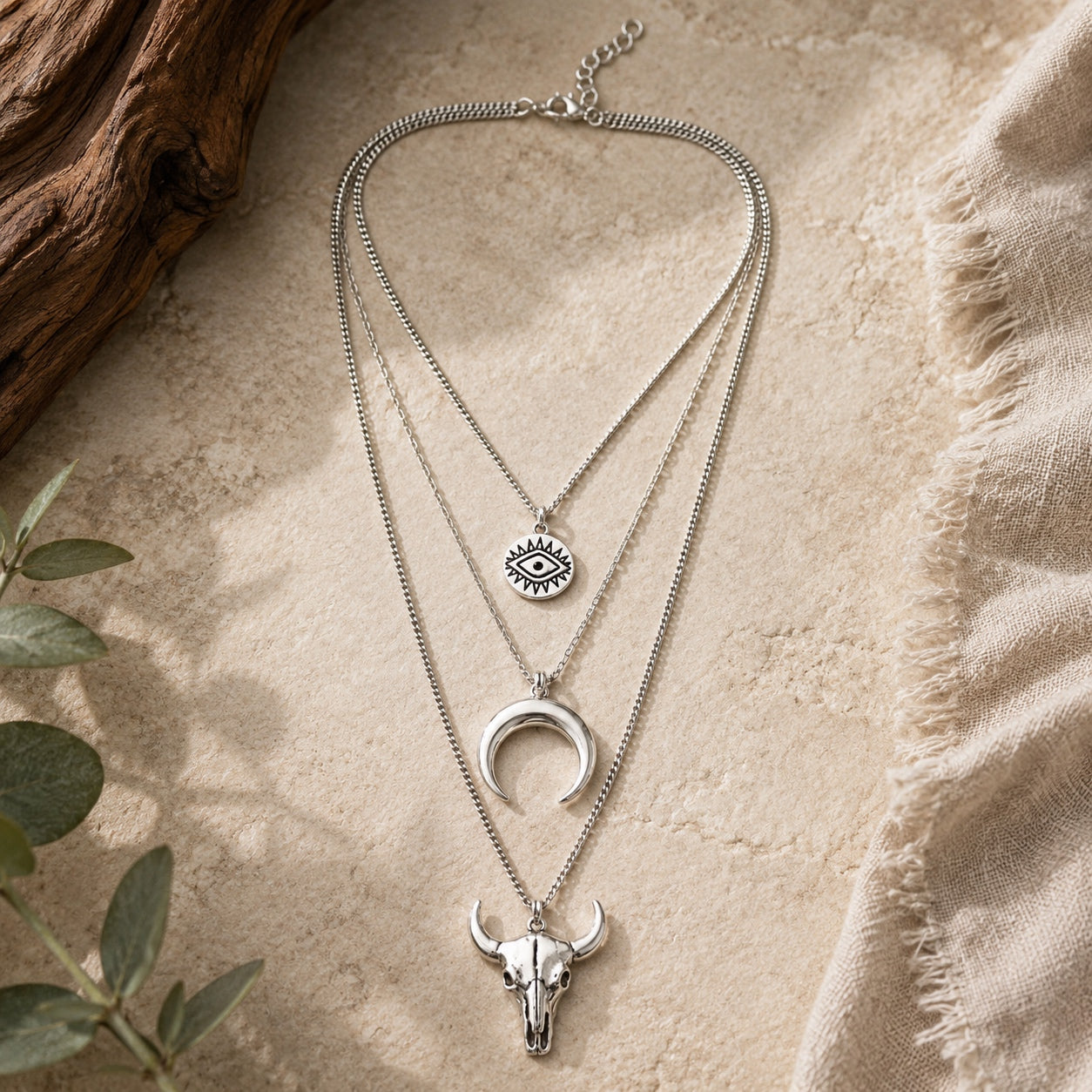 Silver layered boho necklace with bull skull pendant, crescent moon necklace, and evil eye charm displayed on a neutral stone background.